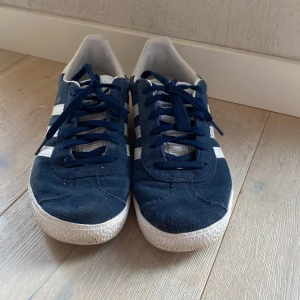 Adidas Gazelle mörkblå sneakers - Säljer ett par klassiska Adidas Gazelle sneakers i mörkblå mocka med vita detaljer och snörning. Skorna har de ikoniska tre ränderna på sidorna och vit sula. Perfekta för en stilren och sportig look.