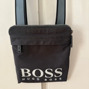 Svart axelväska från Hugo Boss - Snygg svart axelväska från Hugo Boss med stort vitt logotryck framtill. Väskan har justerbart axelband i grått och svart samt dragkedja upptill. Perfekt för att bära med sig det viktigaste på stan eller till skolan.