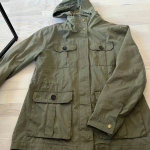 Olivgrön parkajacka från Hampton Republic - Säljer en olivgrön parkajacka från Hampton Republic med huva och flera fickor framtill. Jackan har knappar och dragkedja samt justerbar huva. Perfekt för kyligare dagar och enkel att matcha med olika outfits.