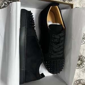 Louboutins skor - Säljer ett par svarta sneakers med mattsvarta nitar på tån och hälen. Skorna har snörning och en klassisk rund tå. Yttermaterialet är mocka och sulan är röd undertill, vilket ger en lyxig känsla.