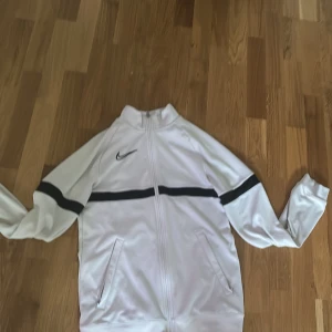 Vit kofta från nike - Säljer en vit träningsjacka från Nike med svart logga och svarta ränder över bröstet och ärmarna. Jackan har dragkedja framtill och två fickor. Tillverkad i Dri-FIT material som andas och passar perfekt till träning.