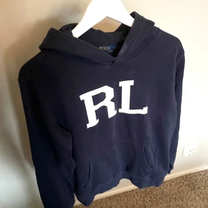 Mörkblå hoodie från Ralph Lauren - Mörkblå hoodie från Polo Ralph Lauren med stor vit RL-logga på bröstet. Tröjan har huva och magficka, perfekt för en avslappnad stil. Klassisk och enkel design som passar till mycket. Inga defekter.