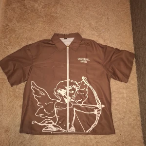 Brun skjorta med ängeltryck från Manfinity - Säljer en brun skjorta från Manfinity med vit dragkedja och tryck av en ängel på framsidan. Skjoryan passar även storlek L. Skjortan har korta ärmar, klassisk krage och texten 'EMOTIONAL ANGEL' på bröstet. Perfekt för dig som vill sticka ut med en unik stil.