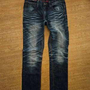Mörkblå jeans från Jacks Denim - Säljer ett par mörkblå jeans från Jacks Denim med snygga slitningar och kontrastsömmar. Modellen är loose fit och har klassiska fem fickor samt läderpatch bak i midjan. Perfekta för en avslappnad stil. 32/32