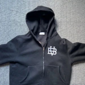 Svart hoodie med dragkedja från BVALDI - Svart hoodie från BVALDI med vit logga på bröstet. Jackan har dragkedja, huva och två fickor framtill. Insidan är fodrad med mjuk fleece för extra komfort och värme. Perfekt för kyliga dagar. Den är lite kort så ifall du är 180 så köp inte rekommenderat är isäfall 170-175