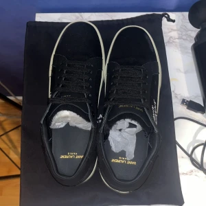 Svarta sneakers från Saint Laurent - Snygga svarta sneakers från Saint Laurent med vit sula och diskret logga på sidan. Skorna har snörning och är tillverkade i textil och skinn. Perfekta för en stilren och modern look.Aldrog använda helt nya äkta skor.