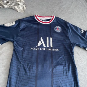PSG fotbollströja Mbappé #7 - Mörkblå Paris Saint-Germain fotbollströja med Mbappé och nummer 7 på ryggen. Tröjan har vita och röda detaljer vid kragen, klubbmärke på bröstet och sponsorloggor. Klassisk design med korta ärmar, perfekt för fans av PSG.