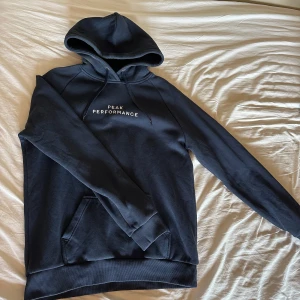 Mörkblå hoodie från Peak Performance - Säljer en mörkblå hoodie från Peak Performance med vit logga på bröstet. Klassisk modell med huva, dragsko och känguruficka. Perfekt för en avslappnad stil och enkel att matcha med jeans eller joggers.