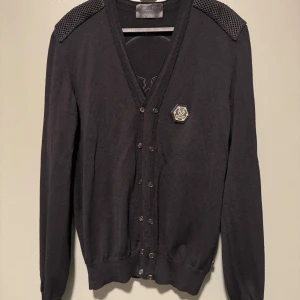 Svart kofta från Philipp Plein med dödskalle - Säljer en svart kofta från Philipp Plein med metallmärke framtill och stickade detaljer på axlarna. Baksidan har en stor dödskalle i struktur. Koftan är tillverkad i mjuk kashmirblandning och har knappar framtill.