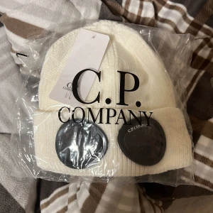  Cp Company mössa vit - Vit ribbstickad mössa från C.P. Company med deras ikoniska svarta glasögondetaljer framtill. Klassisk design med uppvikt kant och tydlig logotyp på glasögonen. Perfekt accessoar för en stilren look.