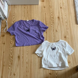 Två croppade t-shirts med fjäril från SHEIN - Säljer två croppade t-shirts från SHEIN, en i lila och en i vit, båda med svart broderad fjärill T-shirtsen har korta ärmar och är ribbade i materialet. Perfekta för en trendig och avslappnad stil. 