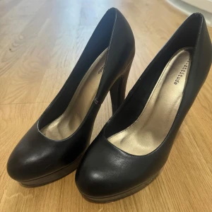 Svarta pumps med klack från Belle - Säljer ett par klassiska svarta pumps från Belle med hög klack och rund tå. Skorna har en plattformsula framtill och är tillverkade i ett slätt material som ger en stilren look. Perfekta för dig som vill ha en tidlös och elegant sko.