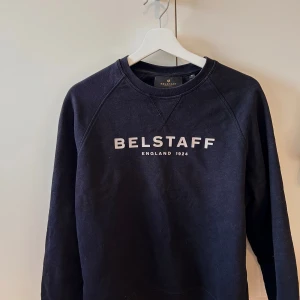 Svart sweatshirt från Belstaff - Svart sweatshirt från Belstaff med vit logga framtill och emblem på ärmen. Tröjan är i 100% bomull och har en klassisk rund halsringning samt långa ärmar. Perfekt för en chill och stilren look.