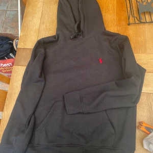 Svart hoodie från Polo Ralph Lauren - Svart hoodie från Polo Ralph Lauren med klassisk röd logga broderad på bröstet. Tröjan har huva med snörning, känguruficka och långa ärmar. Perfekt för dig som gillar stilren och enkel streetwear.
