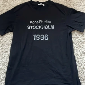 Svart Acne Studios t-shirt 1996 - Svart t-shirt från Acne Studios med tryck i vitt framtill: 'Acne Studios STOCKHOLM 1996'. Klassisk rund hals och korta ärmar. Skön passform och stilren design, perfekt för dig som gillar minimalistiska statement-plagg.