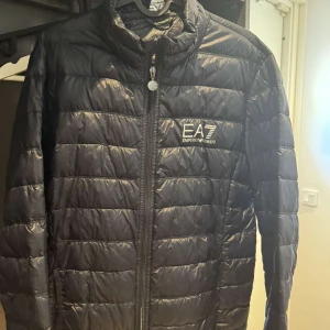 Svart pufferjacka från EA7 Emporio Armani - Säljer en svart pufferjacka från EA7 Emporio Armani med quiltad design och dragkedja framtill. Jackan har hög krage, diskret logga på bröstet och är perfekt för kyliga dagar. Lätt och smidig att bära, passar till de flesta outfits.