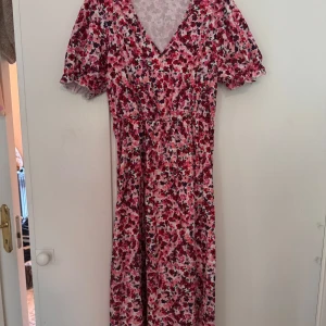 Blommig långklänning med v-ringning - Långklänning med v-ringning och korta puffärmar. Klänningen har ett färgstarkt blommönster i rosa, rött och lila på vit botten. Resår i midjan ger en snygg siluett och materialet känns lätt och luftigt, perfekt för varmare dagar.