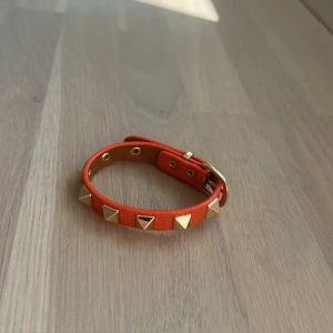 Orange armband med guldnitar - Coolt orange armband i skinn med trekantiga guldnitar runt om. Justerbart spänne i guld och detaljerade öljetter. Perfekt accessoar för att ge din outfit en edgy vibe. VALENTINO LIKNANDE ARMBAND ❤️
