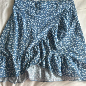 Blommig blå volangkjol från SHEIN - Supersöt blå kjol från SHEIN med vitt blommönster och volangdetaljer. Kjolen har en  dragkedja i sidan för enkel påklädning