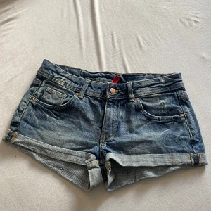 Blå low waist jeansshorts från Divided - Snygga blå jeansshorts från Divided med klassisk femficksdesign och uppvikta benslut. Shortsen har gylf med knapp och dragkedja samt coola slitningar för en avslappnad look. Perfekta för varma dagar och enkel att matcha med allt.