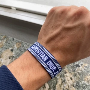 Blått armband Christian Dior J'Adior - Snyggt blått tygarmband från Christian Dior med texten 'CHRISTIAN DIOR J'ADIOR' broderad i vitt. Armbandet har justerbar knytning och fransar i ändarna för en cool och avslappnad look. Perfekt accessoar för att lyfta din outfit.