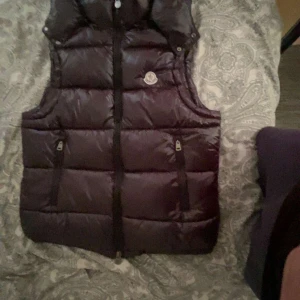 Svart dunväst från Moncler med huva - Säljer en svart dunväst från Moncler med avtagbar huva och två dragkedjefickor framtill. Västen har klassisk quiltad design, blänkande yta och Moncler-logga på bröstet. Perfekt för lager-på-lager och snygg till streetwear eller chill outfits.