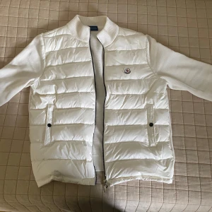 Vit Moncler pufferjacka med stickade ärmar - Säljer en clean vit pufferjacka från Moncler med stickade ärmar och quiltad front. Jackan har dragkedja, två fickor med knappar och broderad Moncler-logga på bröstet.  STORLEK XL, men passar definitivt M och L också. NFC på loggan!