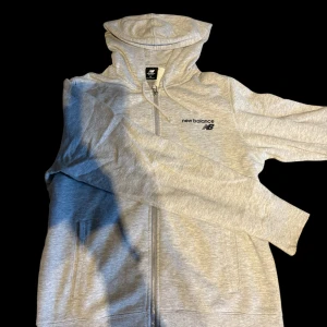 Grå hoodie från New Balance  - Snygg ljusgrå hoodie från New Balance med dragkedja och huva. Tröjan har diskret logga på bröstet, långa ärmar och praktiska fickor framtill. Perfekt för chill eller träning. Tillverkad i mjuk bomullsblandning för extra komfort.