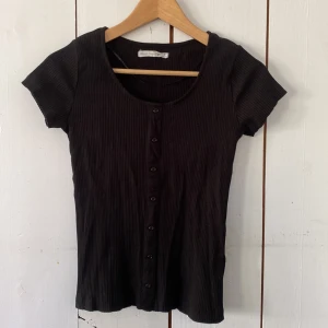 Svart ribbad topp med knappar Black Story - Svart ribbad topp från Black Story med korta ärmar och rund halsringning. Toppen har knappar hela vägen framtill och är gjord i ett mjukt, stretchigt material som sitter snyggt på kroppen. Perfekt till jeans eller kjol för en clean look.