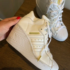 Adidas Superstar vita sneakers med kilklack - Unika Adidas Superstar sneakers i vitt skinn med kilklack och guldiga detaljer. Klassiska tre ränder på sidan, snörning och guldmärkning på plös och sida. Perforerad ovandel och ikonisk snäcktå framtill. Perfekta för dig som vill sticka ut med extra höjd.