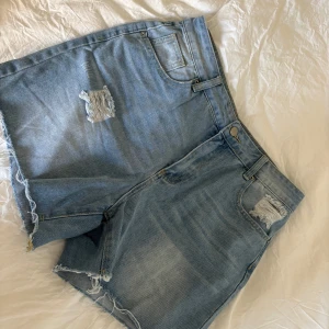 Ljusblå jeansshorts med slitningar - Snygga ljusblå jeansshorts med råa kanter och slitna detaljer framtill. Klassisk femficksmodell med bälteshällor och dragkedja. Perfekta för varma dagar när du vill ha en avslappnad och trendig look. Endast använda en gång i mycket bra skick!