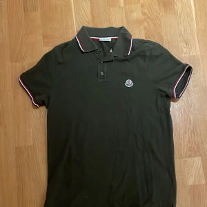 Mörk grön Moncler pikétröja med röd/vit kant - Snygg svart pikétröja från Moncler med klassisk krage och knäppning framtill. Tröjan har röda och vita ränder på krage och ärmslut samt Moncler-logga på bröstet. Perfekt för en stilren och sportig look.