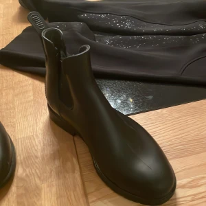 Svarta ridskor i syntet, stl 35 - Svarta ridskor köpta på hööks. De har en del repor här o där och på en sko har tygsidan lossnat från halva bakre delen av skon på den högra sidan av vänster sko. De är rätt gamla och har används rätt mycket vilket också syns en del. Tillverkade i syntetmaterial med dragflik bak för enkel påtagning. Perfekta för dig som vill ha en snygg look till din vanliga ridoutfit. Det dåliga syns inte superväl om man inte kollar lite närmre o att den släppt i tygfästet märks ej om du inte drar lite i tyget.