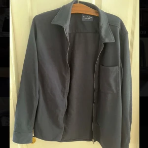 Blå overshirt från Bondelid i M - Stilren svart overshirt från Bondelid med klassisk krage, bröstficka och dragkedja framtill. Perfekt att slänga över en t-shirt för en clean look. Tillverkad i mjuk bomull som känns skön mot huden. Enkel och tidlös design som funkar till det mesta.