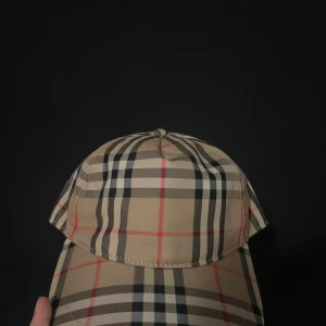 Burberry rutig keps i beige och svart - Snygg keps från Burberry med klassiskt rutigt mönster i beige, svart, vitt och röda linjer. Justerbar baktill med svart spänne och tydlig Burberry London England-patch. Tillverkad i bomull och polyester för en skön passform.