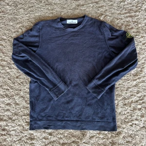 Mörkblå Stone Island sweatshirt - Mörkblå sweatshirt från Stone Island med klassisk rund hals och ikonisk logotyp-patch på vänster ärm. Tröjan har långa ärmar och är tillverkad i mjuk bomull, perfekt för en clean och stilren look.