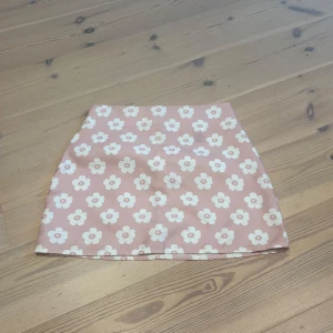 Rosa blommig kortkjol - Supergullig rosa kortkjol med vita blommor över hela tyget. Kjolen har en rak passform och är perfekt för dig som gillar en söt och lekfull stil. Materialet känns mjukt och lätt, perfekt för varmare dagar.