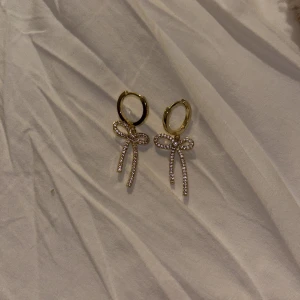 Guldiga örhängen med rosett och strass - Superfina örhängen i guld med hängande rosetter täckta av glittrande strass. Perfekta för att ge din look en söt och trendig touch. Runda hoops med detaljerade rosetter som verkligen sticker ut. Också använt sparsamt då jag inte passar så bra i guld :,) 
