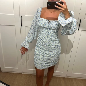 Blommig kortklänning från Adoore - Supersöt kortklänning från Adoore med ljusblått blommönster på vit botten. Klänningen har långa puffärmar, fyrkantig ringning och smickrande rynkade detaljer. Perfekt för sommarens alla tillfällen!