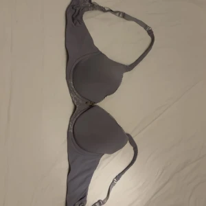 Ljuslila bh med spets och rosett - Supersöt ljuslila bh med vadderade kupor, spetsdetaljer längs kanterna och en liten satinrosett med guldfärgad berlock framtill. Justerbara axelband och hakknäppning bak. Perfekt för dig som gillar en feminin och stilren look. 