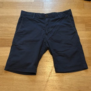 Mörkblå Bowery slim fit shorts - Snygga mörkblå shorts från Bowery i slim fit-modell. Klassisk design med fickor på sidorna och hällor för bälte. Tillverkade i mjuk bomull med lite stretch för extra komfort. Perfekta för varma dagar när du vill ha en clean och stilren look. Det är bara att höra av dig vid minsta lilla fundering😀