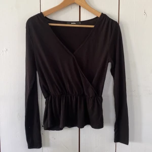 Svart omlottopp från Bik Bok - Svart omlottopp från Bik Bok med v-ringning och markerad midja. Toppen har långa ärmar och en lätt volang nedtill som ger en snygg siluett. Perfekt att styla med jeans eller kjol för en trendig look.
