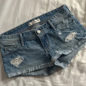 Blå jeansshorts från Hollister - Snygga blå jeansshorts från Hollister med slitna detaljer och råa kanter. Klassisk femficksmodell med låg midja och coola hål med vit spets under. Perfekta för sommaren och ger en avslappnad vibe.