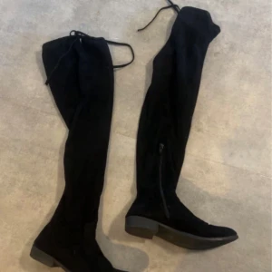 Svarta overknee stövlar i mocka - Snygga svarta overknee stövlar i mjuk mocka med platt sula och rund tå. Stövlarna har snörning upptill och dragkedja på insidan för enkel påtagning. Perfekta för att lyfta vilken outfit som helst under höst och vinter . Storlek 41..