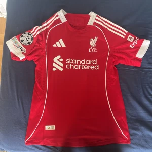Liverpool matchtröja Adidas Wirtz 27 - Liverpool FC matchtröja i rött med vita detaljer, Adidas-logga och klubbmärke på bröstet. Kortärmad modell med tryck 'WIRTZ 27' på ryggen och Premier League-logga. Tillverkad i ventilerande funktionsmaterial, perfekt för fotboll eller supporterliv.