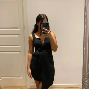 Svart kort klänning med knytband - Snygg svart ärmlös klänning med v-ringning och knytband i midjan. Modellen är kort och har en figurnära passform som framhäver siluetten. Perfekt för dig som gillar stilrena och enkla plagg med en twist.