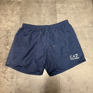 Marinblå badshorts EA7 Emporio Armani - Snygga marinblå badshorts från EA7 Emporio Armani med vit logga på vänster ben. Shortsen har elastisk midja med snörning och sidofickor. Tillverkade i snabbtorkande syntetmaterial och har meshfoder på insidan. Perfekta för stranden eller poolen.