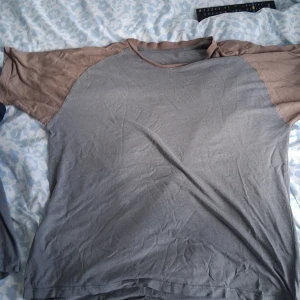 Grå t-shirt med blå färg - T-shirt i avslappnad modell med grå framsida och blå. Rund halsringning med matchande brunt kantband. Perfekt för en chill och enkel stil. Mjuk och skön känsla.