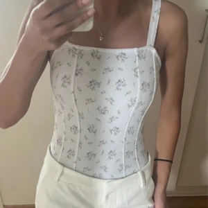 Vit blommig body med smala axelband - Supersöt vit body med små lila och gröna blommor, smala axelband och markerade sömmar framtill. 