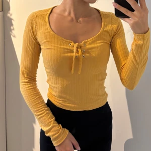 Gul ribbad långärmad topp med snörning - Superfin gul ribbad topp med lång ärm och snygg snörning framtill vid urringningen. Figurnära passform och stretchigt material som sitter skönt. Perfekt för dig som vill sticka ut med färg och coola detaljer.